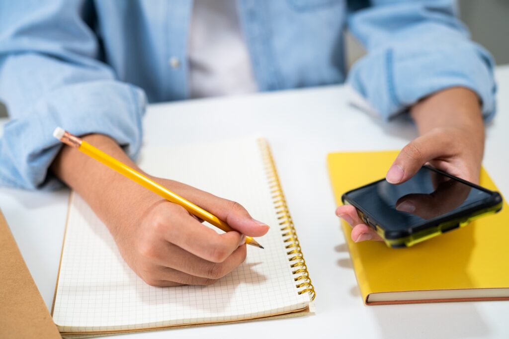 Teenage write in notebook and use mobile phone search data online to online learning university